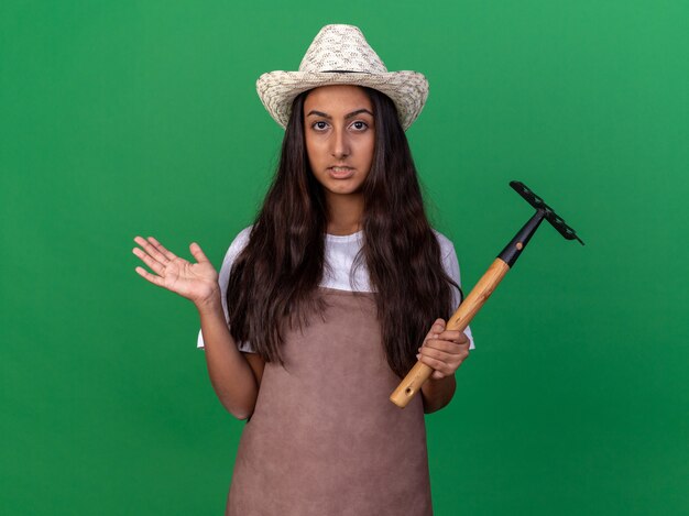 Giovane ragazza del giardiniere in grembiule e cappello estivo che tiene il mini rastrello confuso e incerto che alza il braccio che sta sopra la parete verde