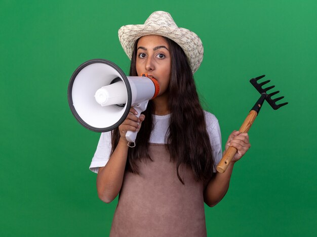 Giovane ragazza del giardiniere in grembiule e cappello estivo che tiene mini rastrello che grida al megafono che sta sopra la parete verde