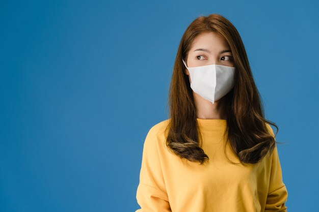 Giovane ragazza dell'Asia che indossa la maschera medica con vestito in un panno casual e guardando uno spazio vuoto isolato su sfondo blu. Autoisolamento, allontanamento sociale, quarantena per la prevenzione del coronavirus