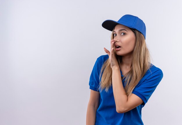 giovane ragazza delle consegne che indossa l'uniforme blu e sussurra berretto isolato sul muro bianco con spazio di copia
