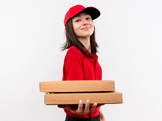 Giovane ragazza delle consegne che indossa l'uniforme rossa e il cappuccio che tengono le scatole per pizza sorridente con la faccia felice in piedi sopra il muro bianco