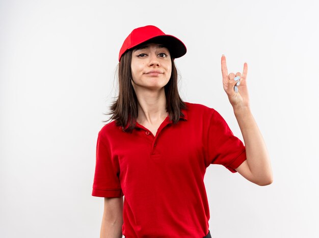 Giovane ragazza delle consegne che indossa l'uniforme rossa e il cappuccio con un sorriso scettico che mostra il simbolo della roccia in piedi sul muro bianco