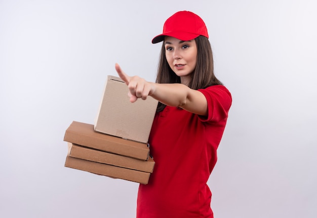 Giovane ragazza delle consegne che indossa la maglietta rossa nel berretto rosso che tiene una scatola e una scatola della pizza e punta le dita di lato su sfondo bianco isolato