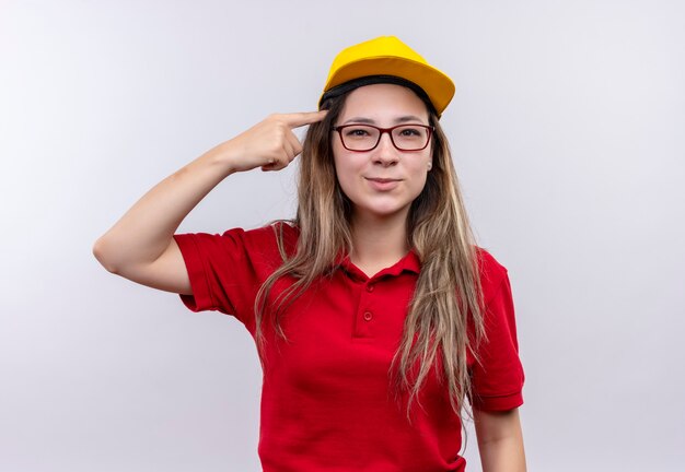 Giovane ragazza delle consegne in maglietta polo rossa e berretto giallo che punta la tempia con il dito indice concentrandosi su un compito