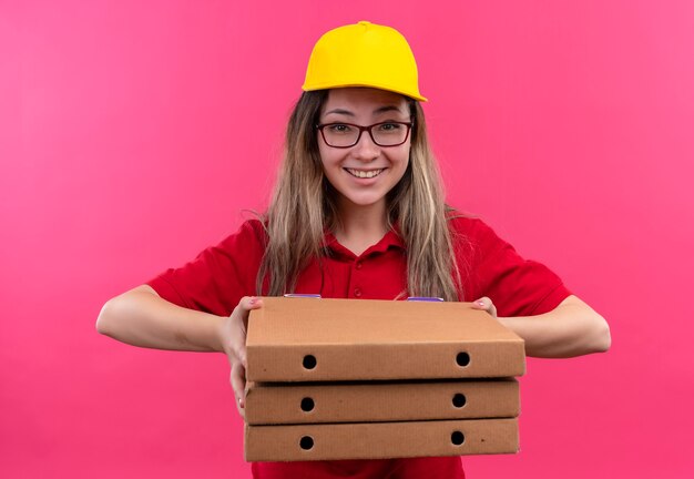 Giovane ragazza delle consegne in maglietta polo rossa e cappuccio giallo che tiene pila di scatole per pizza guardando la telecamera ampiamente sorridente