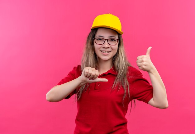 Giovane ragazza delle consegne in polo rossa e cappuccio giallo che punta a se stessa soddisfatto di sé e orgoglioso mostrando i pollici in su sorridente