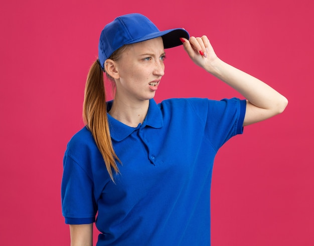 Giovane ragazza delle consegne in uniforme blu e berretto che guarda da parte con un'espressione confusa con la mano sulla testa