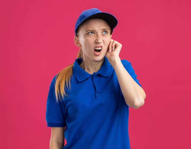 Giovane ragazza delle consegne in uniforme blu e berretto che guarda in alto confusa e dispiaciuta