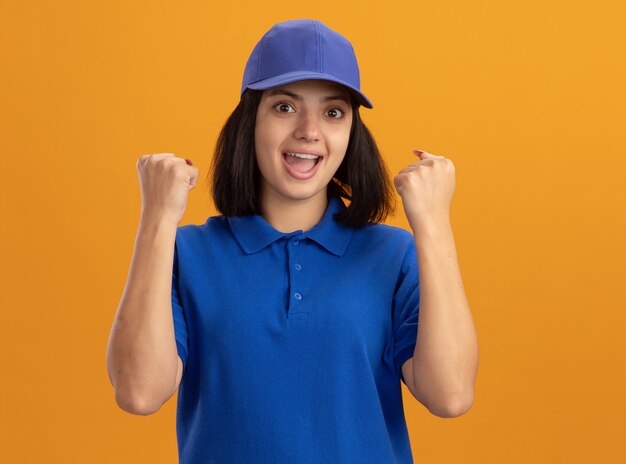 Giovane ragazza delle consegne in uniforme blu e berretto che stringe i pugni felice ed eccitato in piedi sopra la parete arancione