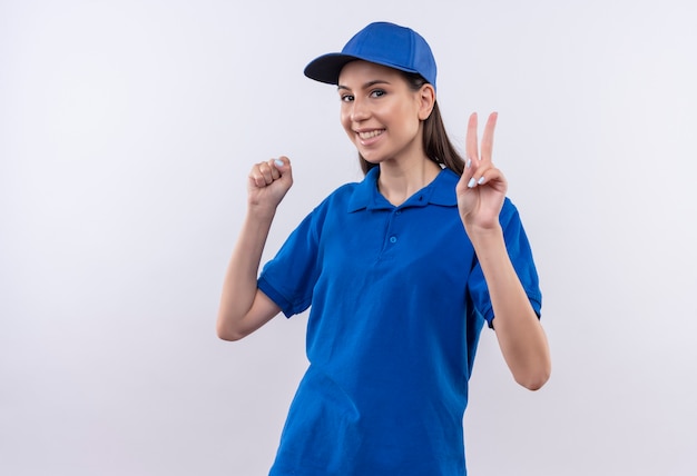 Giovane ragazza delle consegne in uniforme blu e berretto che stringe il pugno che mostra il segno di vittoria