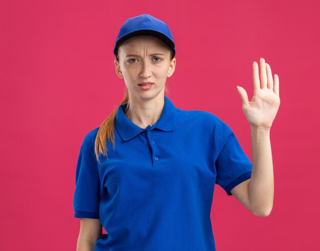 Giovane ragazza delle consegne in uniforme blu e berretto con un'espressione seria e sicura che mostra il braccio aperto