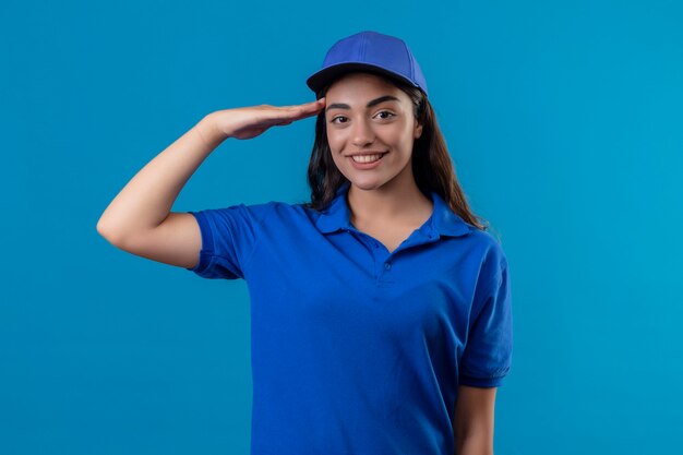 Giovane ragazza delle consegne in uniforme blu e cappello salutando guardando la fotocamera con un sorriso fiducioso sul viso in piedi su sfondo blu