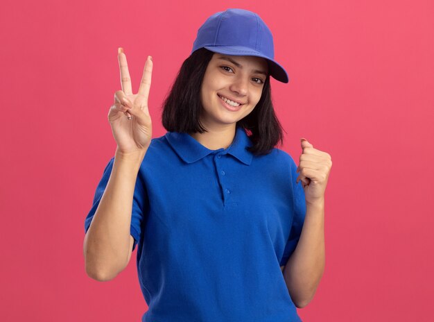 Giovane ragazza delle consegne in uniforme blu e cappello sorridente che mostra il segno di v che stringe il pugno in piedi felice e positivo sopra la parete rosa