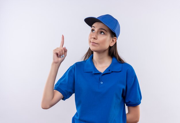 Giovane ragazza delle consegne in uniforme blu e cappuccio che indica con il dito indice in alto con un'espressione seria sicura sul viso