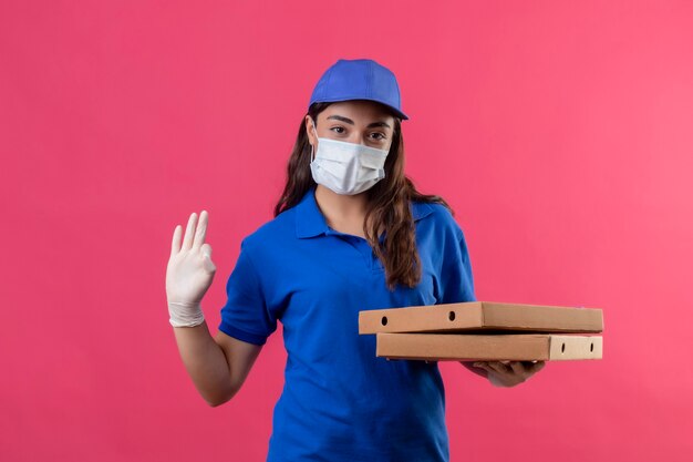 Giovane ragazza delle consegne in uniforme blu e cappuccio che indossa la maschera protettiva per il viso e guanti tenendo le scatole per pizza guardando la telecamera con fiduciosa espressione seria facendo segno ok in piedi su rosa b