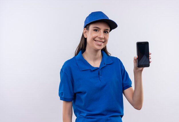 Giovane ragazza delle consegne in uniforme blu e cappuccio che mostra smartphone sorridente ampiamente con la faccia felice