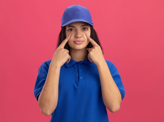 Giovane ragazza delle consegne in uniforme blu e cappuccio che punta con il dito indice ai suoi occhi in piedi sopra il muro rosa