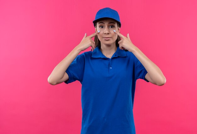 Giovane ragazza delle consegne in uniforme blu e cappuccio che punta con le dita agli occhi