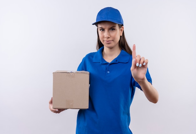 Giovane ragazza delle consegne in uniforme blu e cappuccio che tiene il pacchetto della scatola che mostra l'avvertimento del dito indice con il fronte accigliato