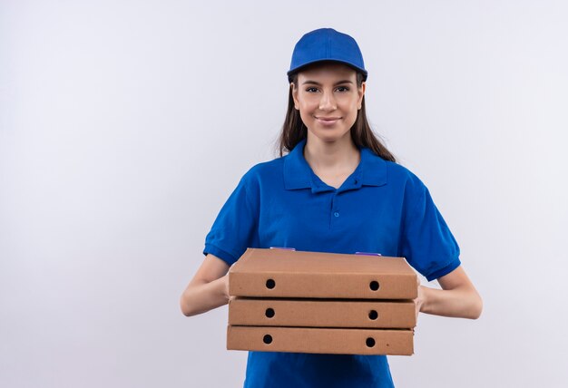 Giovane ragazza delle consegne in uniforme blu e cappuccio che tiene la pila di scatole per pizza che guarda l'obbiettivo con un sorriso fiducioso sul viso