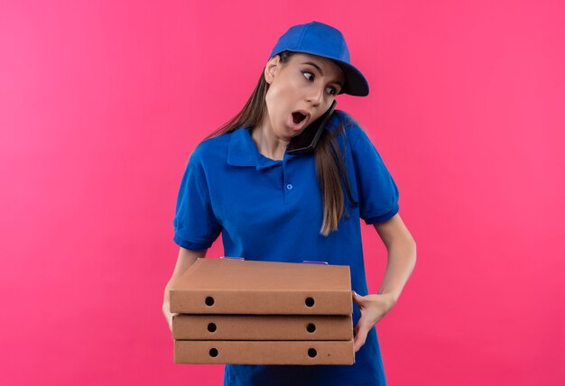 Giovane ragazza delle consegne in uniforme blu e cappuccio che tiene la pila di scatole per pizza che sembra sorpresa mentre parla al telefono cellulare