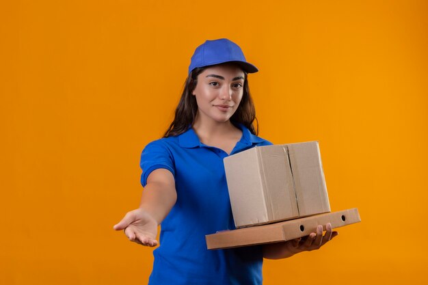 Giovane ragazza delle consegne in uniforme blu e cappuccio che tiene le scatole di cartone cercando fiducioso rendendo il gesto di benvenuto con la mano in piedi su sfondo giallo