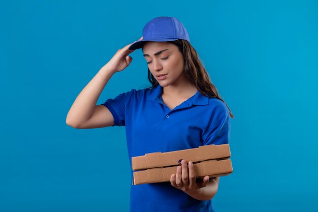 Giovane ragazza delle consegne in uniforme blu e cappuccio che tiene le scatole per pizza alla ricerca di malessere toccando la testa che soffre di mal di testa in piedi su sfondo blu