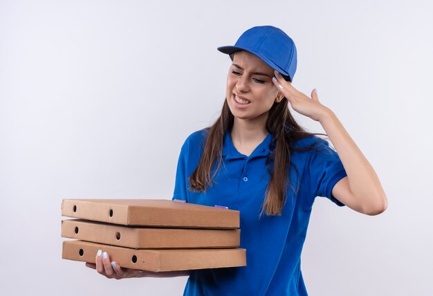 Giovane ragazza delle consegne in uniforme blu e cappuccio che tiene le scatole per pizza cercando dolore alla testa commovente stanco e oberato di lavoro