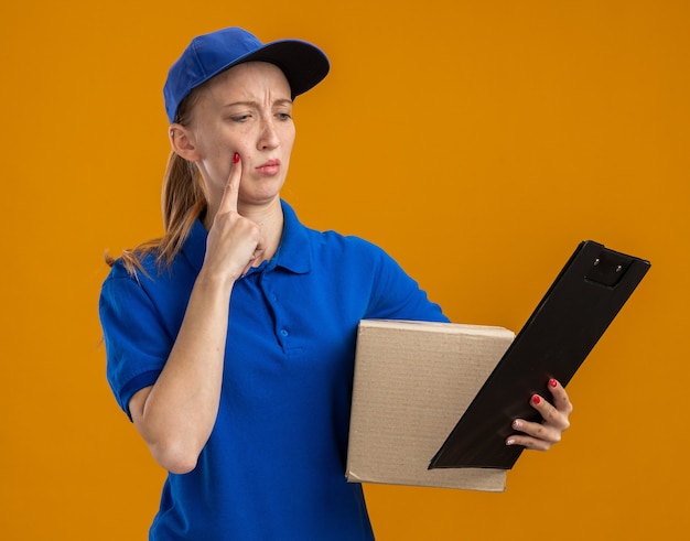 Giovane ragazza delle consegne in uniforme blu e cappuccio che tiene una scatola di cartone e appunti che la guardano con una faccia seria