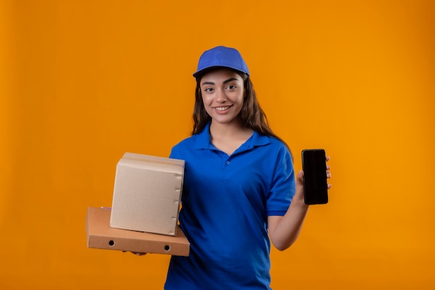 Giovane ragazza delle consegne in uniforme blu e cappuccio tenendo le scatole di cartone che mostra lo smartphone sorridente fiducioso in piedi su sfondo giallo
