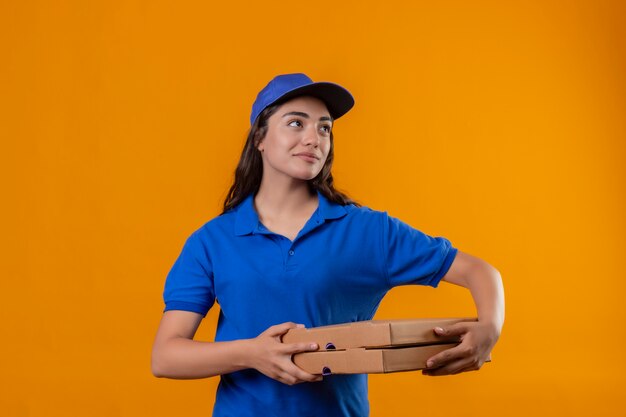 Giovane ragazza delle consegne in uniforme blu e cappuccio tenendo le scatole per pizza guardando da parte con un sorriso fiducioso sul viso in piedi su sfondo giallo