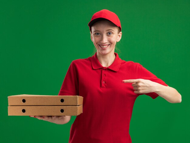 Giovane ragazza delle consegne in uniforme rossa e berretto che tiene in mano scatole di pizza puntate con il dito indice verso di loro sorridendo fiducioso in piedi sul muro verde