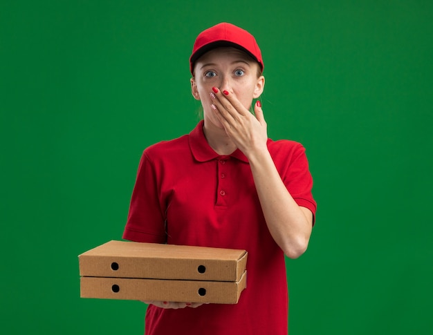 Giovane ragazza delle consegne in uniforme rossa e berretto che tiene in mano scatole di pizza scioccata che copre la bocca con la mano in piedi sul muro verde