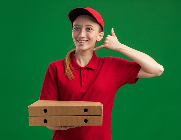 Giovane ragazza delle consegne in uniforme rossa e berretto che tiene in mano scatole per pizza facendomi gesto di chiamarmi sorridente fiducioso in piedi sul muro verde