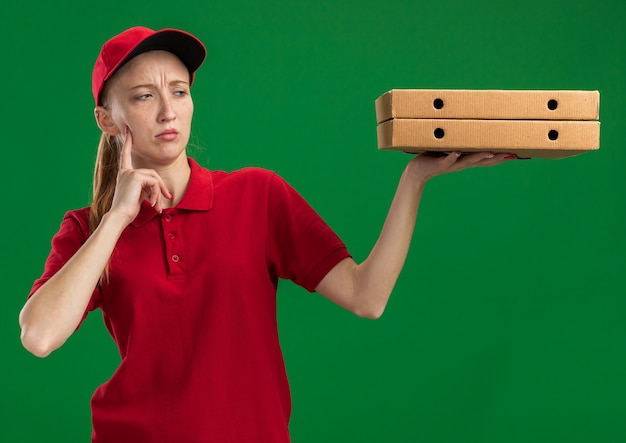 Giovane ragazza delle consegne in uniforme rossa e berretto che tiene in mano scatole per pizza guardandoli perplessi in piedi sul muro verde