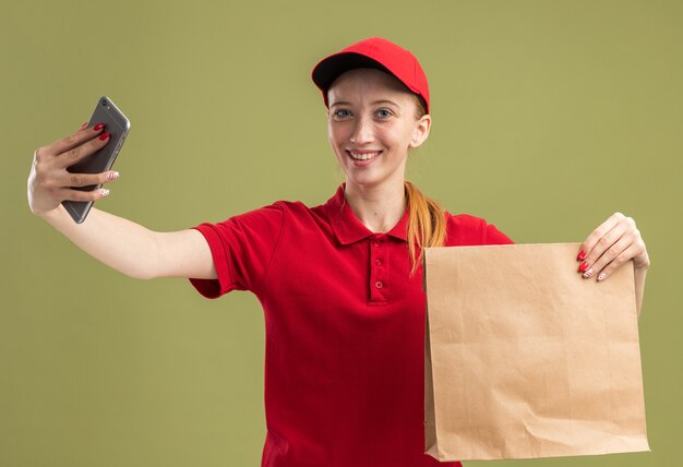 Giovane ragazza delle consegne in uniforme rossa e berretto che tiene un pacchetto di carta facendo selfie utilizzando smartphone sorridendo allegramente sul muro verde