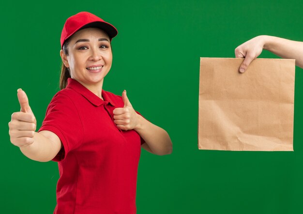 Giovane ragazza delle consegne in uniforme rossa e berretto sorridente che mostra i pollici in su mentre riceve un pacco di carta in piedi sul muro verde