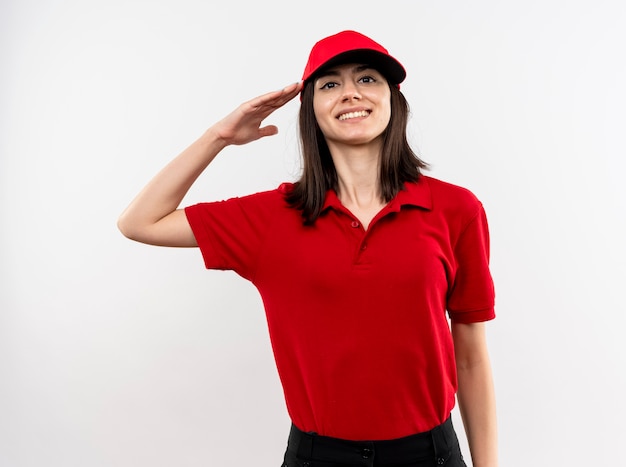 Giovane ragazza di consegna che indossa l'uniforme rossa e berretto che saluta e sorridente fiducioso in piedi sopra il muro bianco