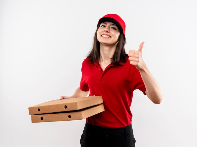 Giovane ragazza di consegna che indossa l'uniforme rossa e cappuccio che tiene le scatole per pizza guardando la fotocamera sorridente con la faccia felice che mostra i pollici in su in piedi su sfondo bianco