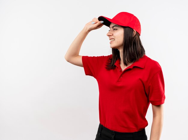Giovane ragazza di consegna che indossa l'uniforme rossa e il cappuccio che osserva da parte essere dispiaciuto di toccare il suo cappuccio in piedi su sfondo bianco