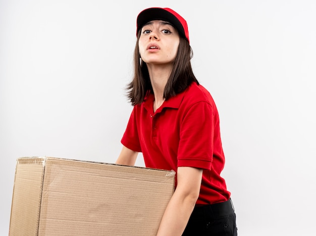 Giovane ragazza di consegna che indossa l'uniforme rossa e il cappuccio che tiene grande scatola di cartone pesante che sembra stanco in piedi sopra il muro bianco
