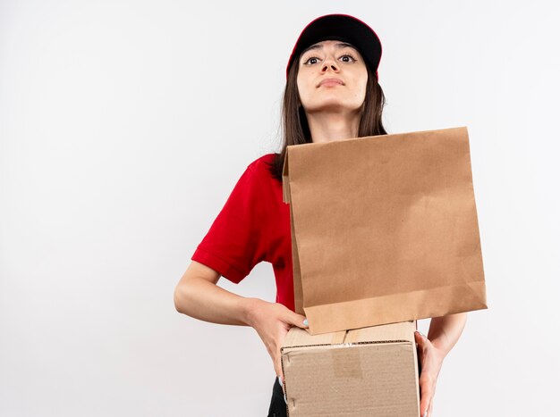Giovane ragazza di consegna che indossa l'uniforme rossa e il cappuccio che tiene il pacchetto di carta e la scatola di cartone che guarda l'obbiettivo con espressione sicura che sta sopra fondo bianco