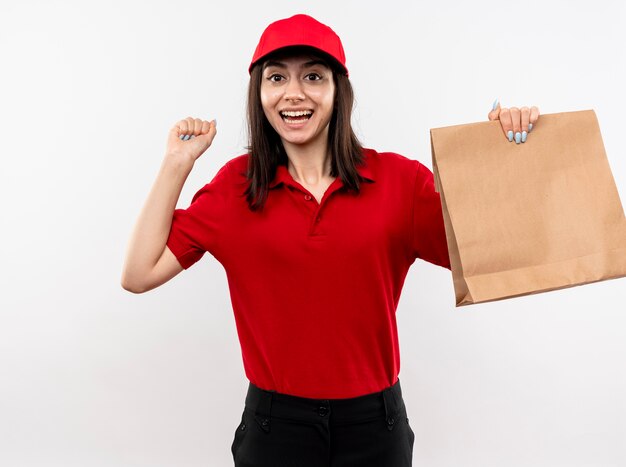 Giovane ragazza di consegna che indossa l'uniforme rossa e la protezione che tiene il pugno di serraggio del pacchetto di carta felice ed eccitato che sta sopra fondo bianco