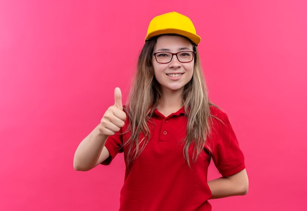 Giovane ragazza di consegna in camicia di polo rossa e berretto giallo sorridente che mostra i pollici in su