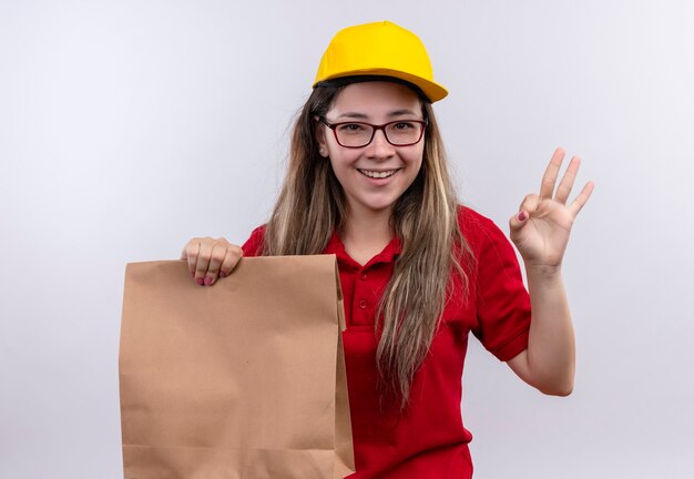 Giovane ragazza di consegna in camicia di polo rossa e cappuccio giallo che tiene il pacchetto di carta che sorride allegramente mostrando segno giusto