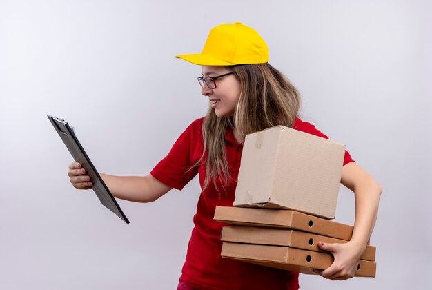 Giovane ragazza di consegna in maglietta di polo rossa e cappuccio giallo che tiene pila di scatole per pizza guardando appunti nell'altra mano cercando confuso