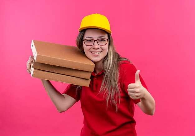 Giovane ragazza di consegna in polo rossa e cappuccio giallo che tiene pila di scatole per pizza guardando la telecamera sorridendo allegramente mostrando i pollici in su