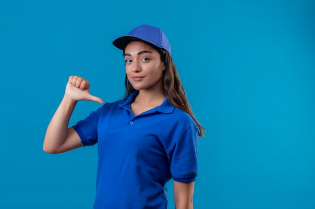 Giovane ragazza di consegna in uniforme blu e berretto cercando fiducioso che punta a se stessa con il pollice soddisfatto di sé e orgoglioso in piedi su sfondo blu