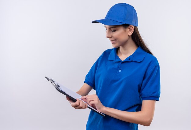 Giovane ragazza di consegna in uniforme blu e berretto guardando le pagine vuote negli appunti con il sorriso sul viso
