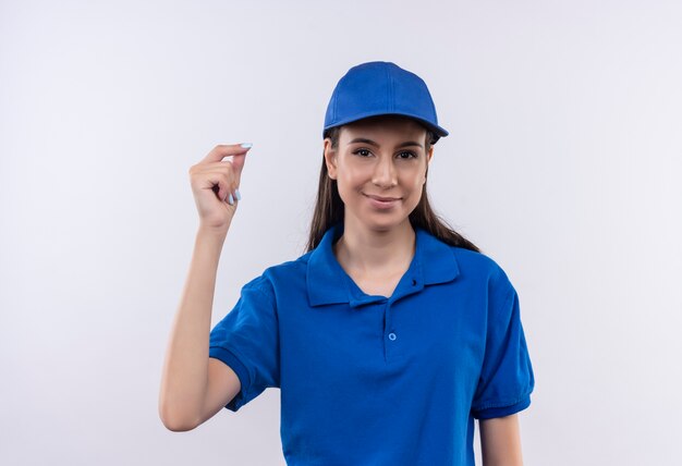 Giovane ragazza di consegna in uniforme blu e cappuccio che fa gesto di soldi sfregando le dita con il sorriso sul viso